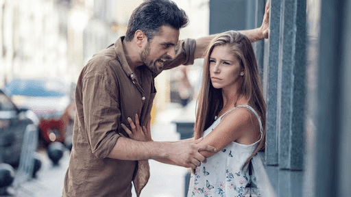 Een man confronteert een vrouw op indringende wijze tegen een muur, wat een voorbeeld toont van misplaatste dominantie. Deze scène benadrukt het verschil tussen agressie en echte, veilige mannelijke kracht.