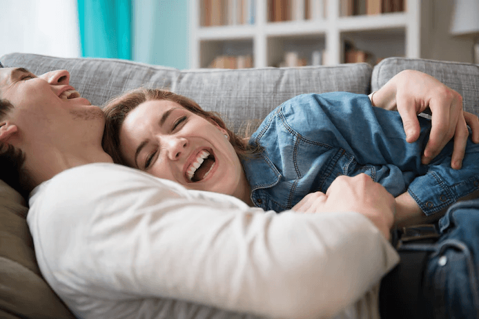 Een jong stel lacht samen op de bank, een moment van intense verbondenheid dat de krachtige hormonale roes weerspiegelt waar mensen soms letterlijk verslaafd aan kunnen raken in de beginfase van verliefdheid.