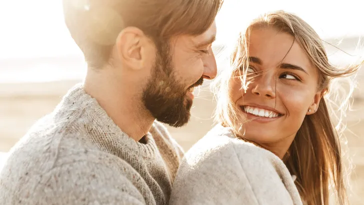 Een jong stel kijkt elkaar liefdevol aan terwijl ze buiten in het zonlicht zitten. Beiden dragen warme, beige truien en glimlachen naar elkaar, wat een gevoel van geluk en verbondenheid uitstraalt. De zon schijnt op de achtergrond, wat zorgt voor een zachte, romantische sfeer.