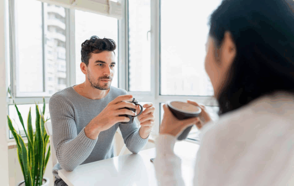 Een man en een vrouw zitten tegenover elkaar aan een tafel met een kop koffie in de hand. De man kijkt de vrouw recht aan, terwijl zij haar blik lijkt af te wenden. De setting is licht en open, maar de lichaamstaal van de vrouw straalt onzekerheid uit — passend bij het gevoel: "Ik ben bang voor oogcontact." Het beeld toont een subtiel spanningsveld in een ogenschijnlijk ontspannen gesprek.