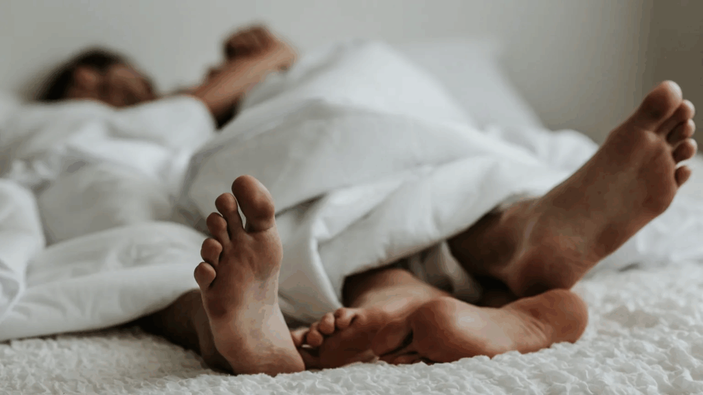 Twee mensen liggen samen in bed onder een wit dekbed, met alleen hun blote voeten zichtbaar. Ondanks de fysieke nabijheid straalt het beeld een zekere afstand of stilte uit. De sfeer roept het gevoel op van: "Er is geen seksuele energie." Het beeld symboliseert gemis aan passie of connectie in een intieme setting.