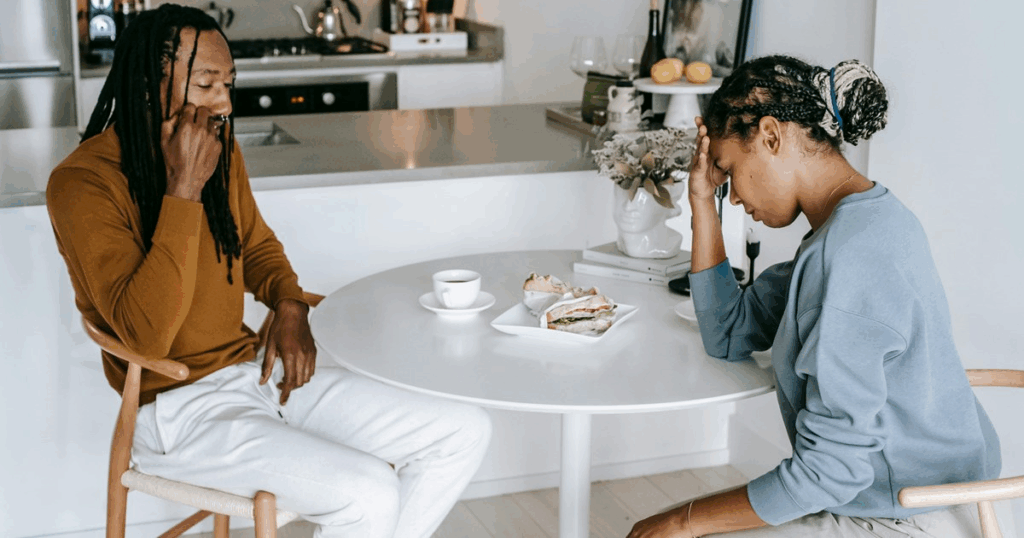 Een man en vrouw zitten aan een ronde tafel met eten voor zich, maar beiden kijken weg en houden hun hoofd vast, zichtbaar gespannen of verdrietig. Ondanks de nabijheid is er geen oogcontact of verbinding, wat een gevoel van "afstand in elk gesprek" oproept. Het beeld weerspiegelt emotionele verwijdering en moeilijkheden in communicatie.