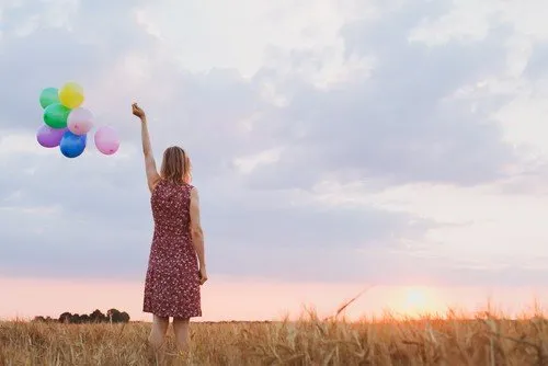Een vrouw in een rode jurk met bloemenpatroon staat in een graanveld bij zonsondergang en houdt een bos gekleurde ballonnen omhoog. Ze kijkt naar de horizon onder een deels bewolkte lucht, wat een gevoel van vrijheid en hoop uitstraalt.