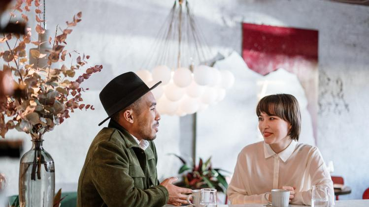 Twee mensen zitten samen aan een tafel in een modern café. De man draagt een groene jas en een zwarte hoed, en praat enthousiast, terwijl de vrouw in een witte blouse glimlachend naar hem luistert. Op tafel staan koffiekopjes en glazen water, en op de achtergrond hangt een stijlvolle lamp met ronde bollen. De sfeer oogt ontspannen en gezellig.