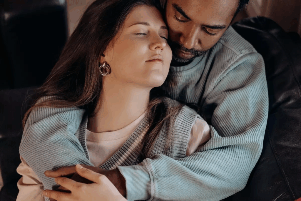 Een vrouw zit met gesloten ogen in de armen van een man, zichtbaar ontspannen en op haar gemak. Zijn omhelzing straalt warmte en bescherming uit, wat een gevoel van veiligheid en geborgenheid oproept. Het moment toont diepe verbondenheid en vertrouwen tussen hen.