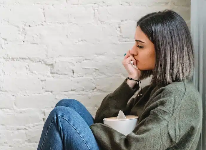 Een vrouw zit met opgetrokken knieën tegen een muur, verdiept in haar gedachten en met een verdrietige blik. Ze houdt een doos tissues vast, alsof ze net gehuild heeft. Haar houding en gezichtsuitdrukking stralen uit: "Ik voel me niet begeerd." Het beeld toont een moment van kwetsbaarheid, gemis en emotionele eenzaamheid.
