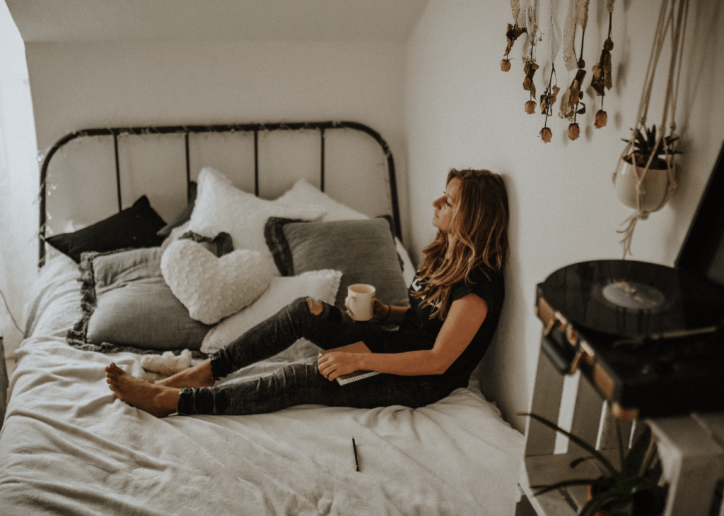 Een vrouw zit dromerig op haar bed, met een kopje in haar hand en een notitieboekje op haar schoot. Ze staart voor zich uit, alsof ze in gedachten verzonken is. De sfeer is rustig maar melancholisch. Haar houding en blik zeggen zonder woorden: "Waarom val ik steeds op onbereikbare mannen?" Het beeld ademt verlangen, reflectie en emotionele afstand.